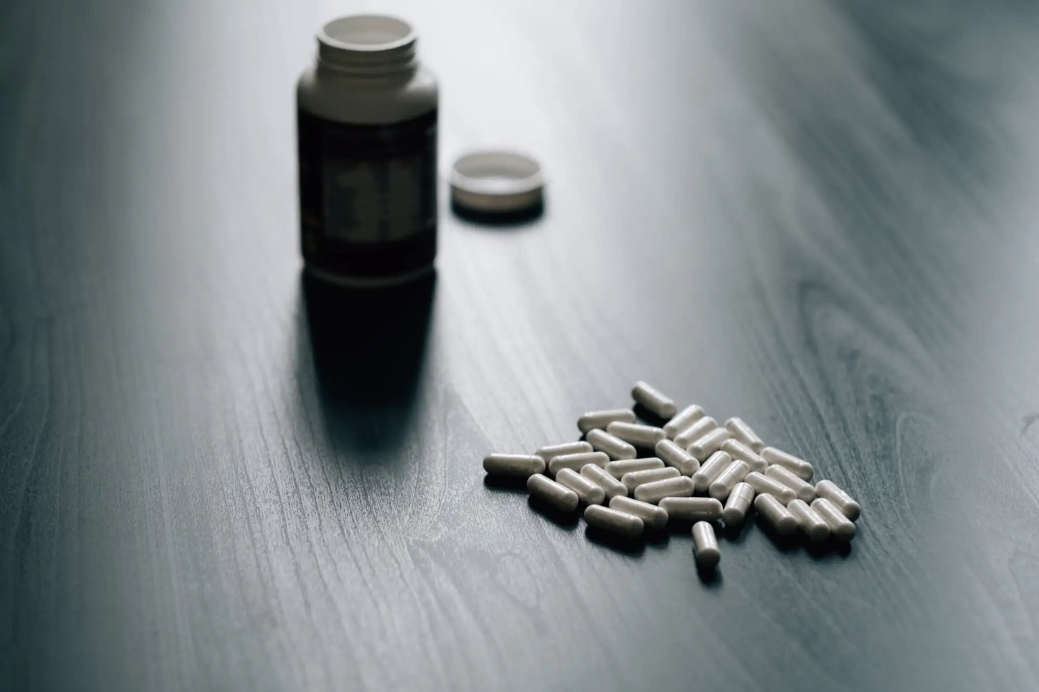 White capsules scattered on a dark wooden surface next to an open, unlabeled pill bottle.